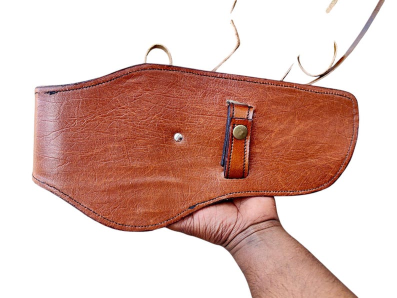 May include: A brown leather holster with a textured surface. The holster features a strap with a brass button closure and a silver rivet. The item is designed to be worn on the waist.
