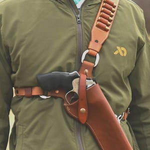 May include: A brown leather shoulder holster and belt, holding a black revolver. The shoulder strap has loops with bullets. The person is wearing a green jacket. The holster is made of leather.