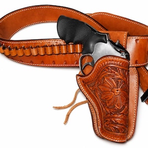 Op de afbeelding: Een lichtbruine lederen holster met gedetailleerde bloemmotieven en twee lederen veters, bevestigd aan een bijpassende lederen patroongordel met munitielussen en een zwarte metalen gesp, waarin een zilverkleurige en zwarte revolver rust.