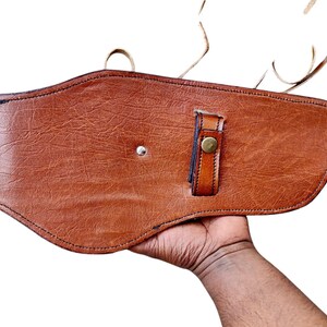 May include: A brown leather holster with a textured surface. The holster features a strap with a brass button closure and a silver rivet. The item is designed to be worn on the waist.