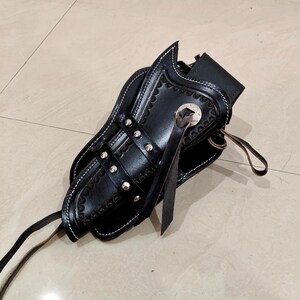 Peut inclure: Un &eacute;tui &agrave; pistolet en cuir noir avec des coutures blanches et des rivets argent&eacute;s. L'&eacute;tui pr&eacute;sente des bords festonn&eacute;s d&eacute;coratifs et un embl&egrave;me en forme d'&eacute;toile. Une sangle et une attache en cuir sont &eacute;galement visibles.