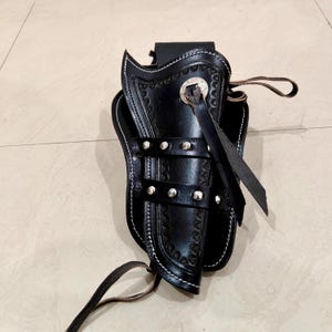 Peut inclure: Un &eacute;tui en cuir noir avec des coutures d&eacute;coratives et des rivets argent&eacute;s. L'&eacute;tui pr&eacute;sente un motif en relief d&eacute;taill&eacute; et une lani&egrave;re en cuir. Les bords sont finis avec des coutures blanches. Un accessoire classique pour porter une arme &agrave; feu.