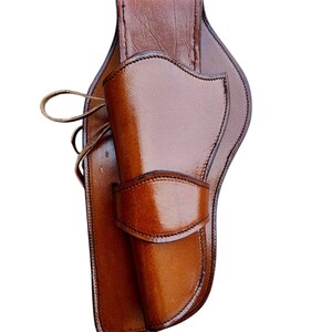 May include: A brown leather holster with a glossy finish. The holster features a curved design with stitched edges and a flap to secure a firearm. A leather strap is visible on the side.