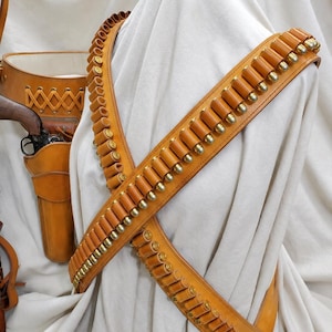 Peut inclure: Un baudrier en cuir beige composé de deux sangles croisées avec des boucles individuelles contenant des cartouches à douille en laiton. Un étui en cuir beige assorti avec un laçage décoratif et une arme à feu est placé sur le côté.