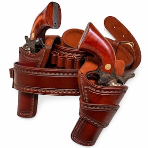 May include: A brown leather double holster gun belt featuring two revolvers with polished wooden grips. The belt includes integrated cartridge loops and decorative scalloped edge stitching, designed to hold two firearms securely in place.