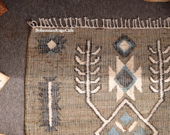 May include: A woven area rug featuring a tan base with geometric patterns in white, dark gray, and light blue. The rug has a fringed edge at the top and includes the text BohemianRugzCafe printed in white on the upper left side of the fabric.