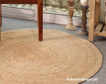 May include: A round, natural-colored jute rug with a braided, circular weave pattern. The rug is positioned on a gray floor beneath a wooden chair. The text "BohemianRugzCafe" is printed in white in the lower right corner of the image.