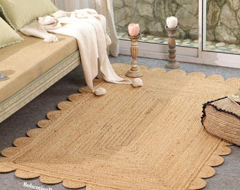 May include: A rectangular area rug made of woven natural tan jute fibres, featuring a decorative scalloped edge border. The rug has a flat, braided texture and is positioned on a grey floor. The text BohemianRugzCafe is visible.