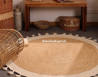 May include: A round, woven jute rug featuring a natural tan center and a wide, off-white border. The edge is decorated with small, evenly spaced tassels. The text "BohemianRugzCafe" is printed in white across the center of the rug.