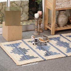 May include: A rectangular woven area rug featuring a natural beige base with repeating blue geometric patterns. The rug has a textured surface and is positioned on a gray floor. The text "BohemianRugzCafe" is printed in white on the rug.
