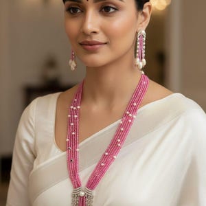 May include: A woman wearing a pink beaded necklace with a silver pendant and matching earrings. The necklace has multiple strands of pink beads interspersed with white pearls. The earrings are long and feature a similar design. The woman is wearing a white top.