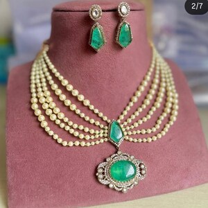 May include: A luxurious jewelry set featuring a multi-strand pearl necklace with a large emerald pendant and matching emerald earrings. The pendant and earrings are accented with small, sparkling stones. The jewelry is displayed on a pink velvet surface.
