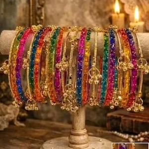 Puede incluir: Una exhibición de pulseras coloridas en una variedad de colores, incluyendo rojo, naranja, amarillo, verde, azul y morado. Las pulseras están adornadas con detalles dorados y pequeños dijes colgantes. Se exhiben en un soporte de madera.