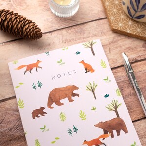 May include: A pink notebook with a woodland theme. The cover features illustrations of bears, foxes, and trees. The word "NOTES" is printed in black on the front of the notebook.