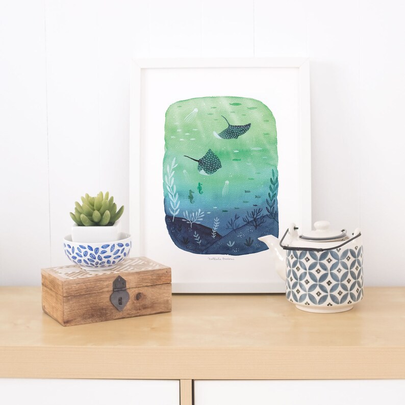 May include: A watercolor painting of a blue and green underwater scene with two stingrays swimming. The painting is framed in a white frame and is sitting on a wooden shelf with a small potted plant and a blue and white teapot.
