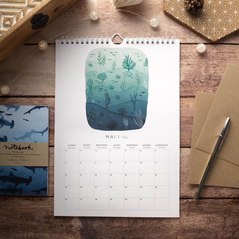 May include: A wall calendar with a watercolor illustration of an underwater scene. The illustration features a variety of sea life, including fish, coral, and seaweed. The calendar is for the month of May and is printed in French and English. The calendar is hanging on a wooden surface.