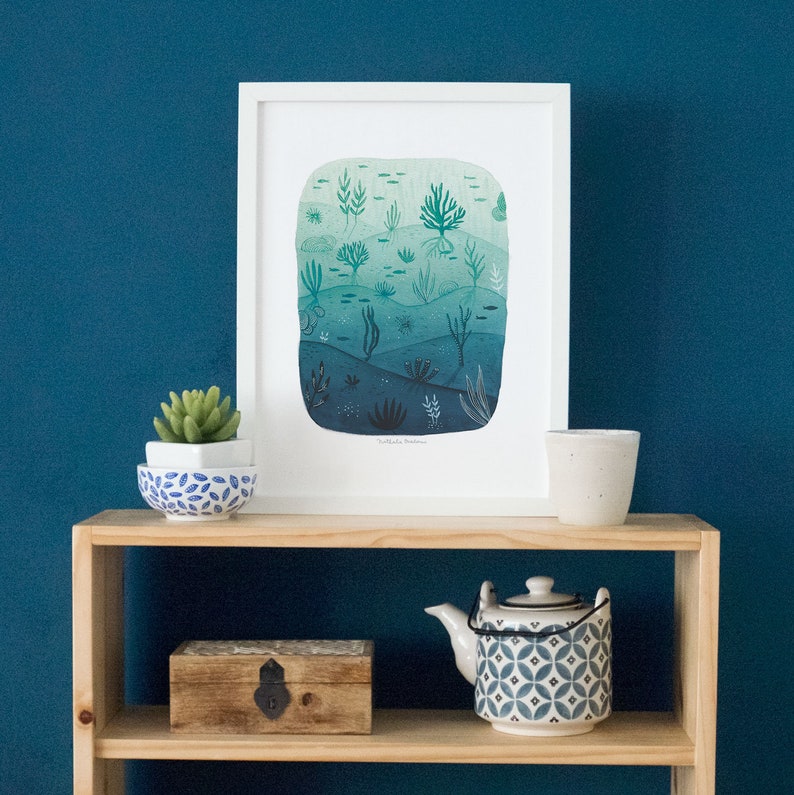 May include: A white framed print of a watercolor illustration of an underwater scene with various shades of blue and green. The print features a variety of sea life, including fish, seaweed, and coral. The text "Subtle Sea Dreams" is printed at the bottom of the print.
