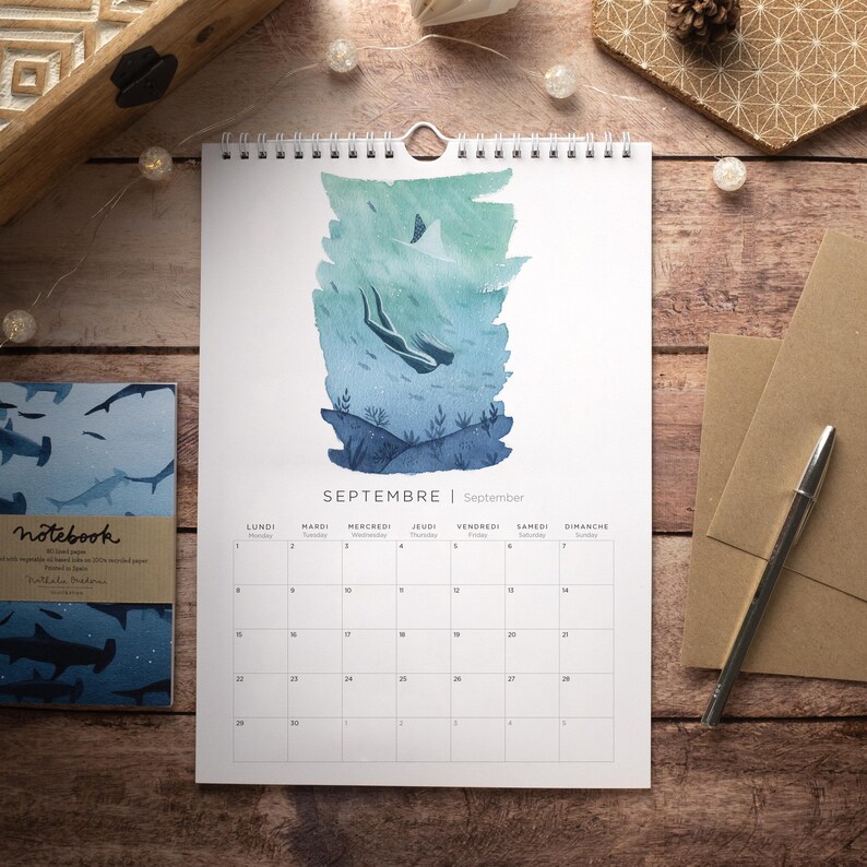 May include: A watercolor calendar featuring a blue and green illustration of a diver in the ocean. The calendar is for September and has the months written in French and English. The calendar is hanging on a wooden surface.