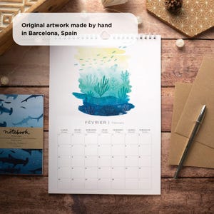 May include: A watercolor illustration of an underwater scene with coral reefs, fish, and a jellyfish. The illustration is on a white calendar with the month of February in French and English. The calendar is on a wooden surface.