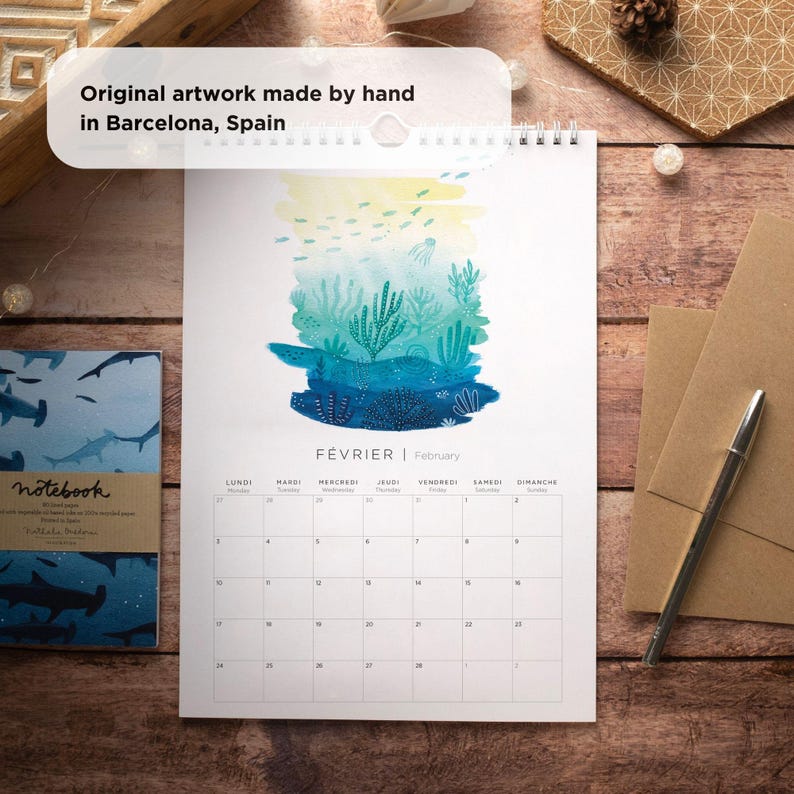 May include: A watercolor illustration of an underwater scene with coral reefs, fish, and a jellyfish. The illustration is on a white calendar with the month of February in French and English. The calendar is on a wooden surface.