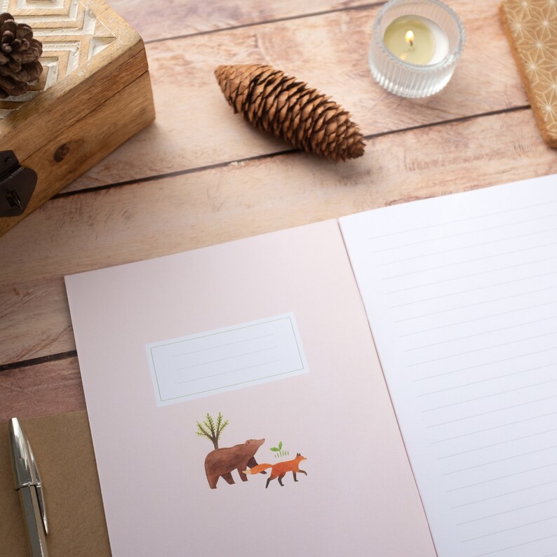 May include: A pink notebook with a brown bear and two foxes on the cover. The notebook has a blank label for writing a name. The notebook is open to a blank page with lined paper.