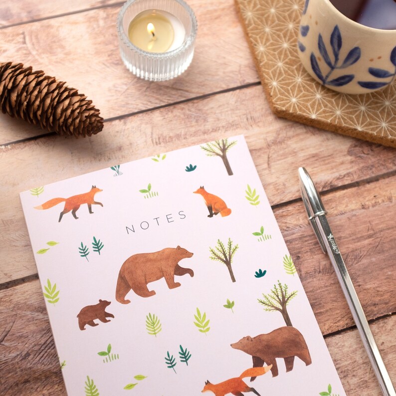 May include: A pink notebook with a woodland theme. The cover features illustrations of bears, foxes, and trees. The word "NOTES" is printed in black on the front of the notebook.