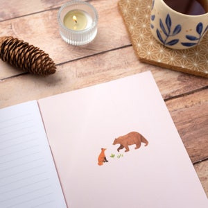 May include: A pink notebook with a lined page on the left and a brown bear and orange fox illustration on the right. The illustration shows the bear and fox facing each other.