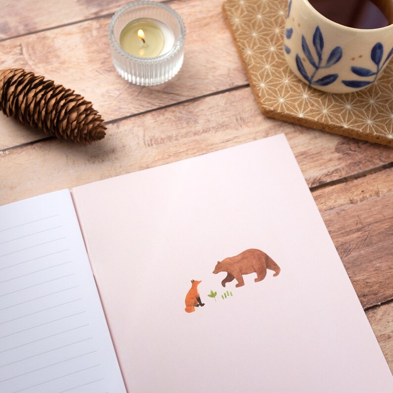 May include: A pink notebook with a lined page on the left and a brown bear and orange fox illustration on the right. The illustration shows the bear and fox facing each other.