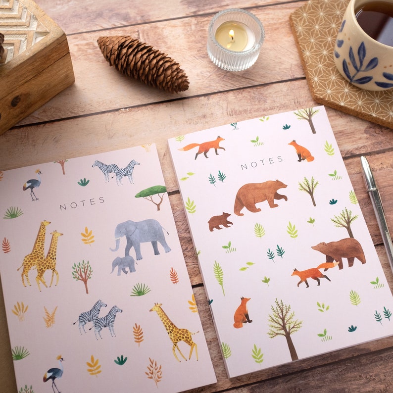 May include: Two pink notebooks with a "NOTES" title and a variety of animal illustrations. One notebook features giraffes, elephants, zebras, and birds. The other notebook features bears, foxes, and trees.