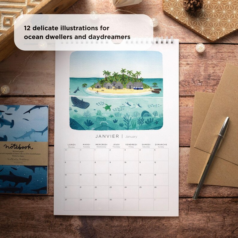 May include: A wall calendar with a watercolor illustration of an island with palm trees and a turquoise ocean. The calendar is open to January and features the French words "JANVIER" and "LUNDI" through "DIMANCHE".