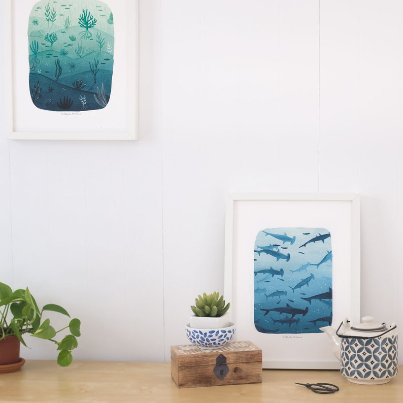 May include: Two framed illustrations of underwater scenes. The top illustration shows a coral reef with a gradient of blue and green colors. The bottom illustration shows a school of hammerhead sharks swimming in a gradient of blue colors. Both illustrations have white frames and are displayed on a white wall.