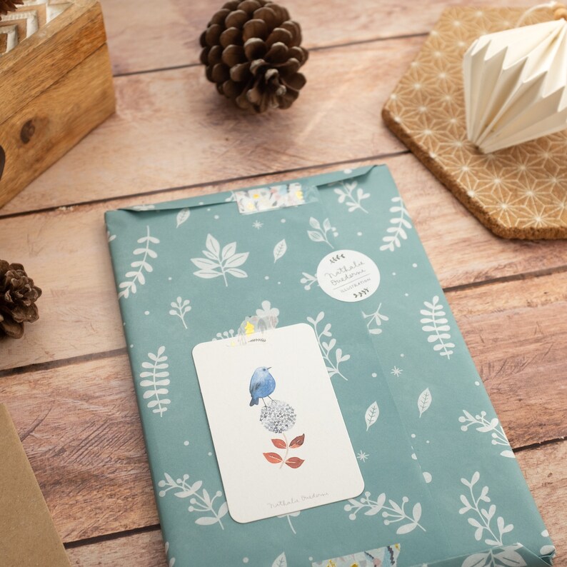 May include: A blue bird perched on a white flower with red leaves on a white card with the text "Nathalie Bussien" on a teal blue wrapping paper with a white floral pattern. The wrapping paper has a round sticker with the text "Nathalie Bussien" on it.