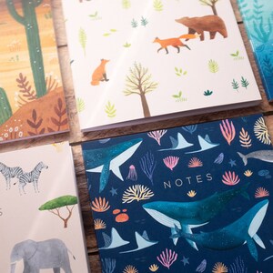 May include: Four different notebooks with colorful illustrations. The first notebook has a desert scene with a cactus and a fox. The second notebook has a forest scene with a bear and foxes. The third notebook has a whale and coral reef scene. The fourth notebook has a zebra and elephant scene. All notebooks have the word "NOTES" printed on the front.