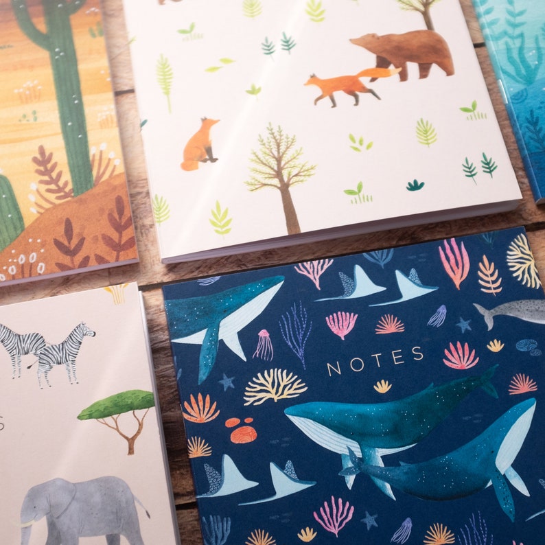 May include: Four different notebooks with colorful illustrations. The first notebook has a desert scene with a cactus and a fox. The second notebook has a forest scene with a bear and foxes. The third notebook has a whale and coral reef scene. The fourth notebook has a zebra and elephant scene. All notebooks have the word "NOTES" printed on the front.