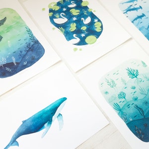May include: Set of four watercolor prints featuring nature scenes. The prints depict a whale, swans in a pond, a underwater scene with coral, and a forest scene with trees and leaves.