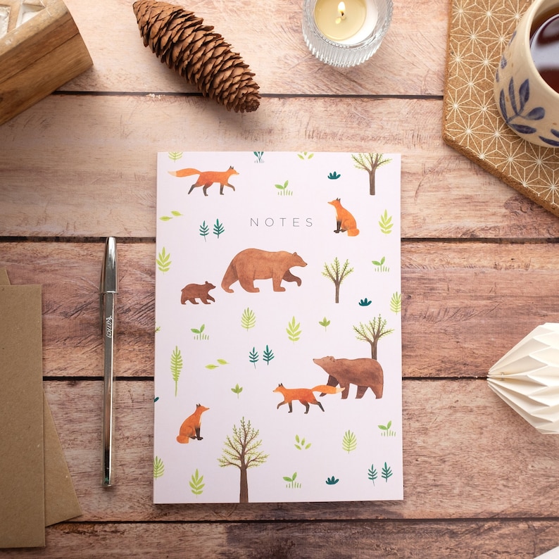 May include: A pink notebook with a woodland theme. The cover features bears, foxes, and trees in a repeating pattern. The word "NOTES" is printed in black at the top of the notebook.