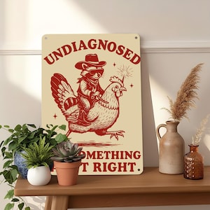 May include: A cream-colored metal sign with the words "UNDIAGNOSED" and "SOMETHING AIN'T RIGHT" in red. The sign features a raccoon wearing a cowboy hat and sunglasses riding a chicken, with a sparkler above the chicken's head.