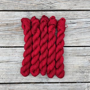 Puede incluir: Cinco madejas de hilo rojo vibrante se disponen sobre una superficie de madera envejecida. El hilo está bien enrollado, mostrando un color rico y saturado. Las tablas de madera proporcionan un fondo rústico, resaltando la textura y el tono del hilo. Ideal para proyectos de manualidades.