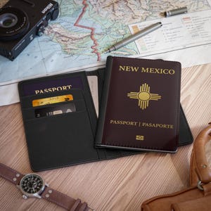 Op de afbeelding: Een zwarte paspoortportemonnee met een New Mexico paspoort en een paspoort erin. Een zwarte Nikon camera, een pen en een horloge met een bruine leren band zijn ook zichtbaar op een houten oppervlak met een kaart.