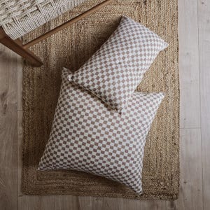 Könnte beinhalten: Zwei dekorative Kissen mit braun-weißem Karomuster liegen auf einem gewebten Jute-Teppich. Ein rechteckiges Kissen liegt auf einem quadratischen Kissen. Die Kissen liegen auf einem Holzboden, ein Teil eines Holzstuhls ist sichtbar.