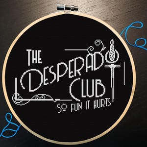 May include: A circular wooden embroidery hoop containing black fabric with white cross-stitched text that reads THE DESPERADO CLUB SO FUN IT HURTS. The design includes a stylized sword graphic on the right side and decorative line accents.