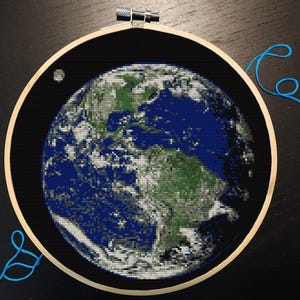 以下が含まれることがあります： 地球のクロスステッチ刺繍。木製のフレームに収められています。青、緑、白、グレーの色合いで詳細にステッチされた大陸が特徴です。左上に小さな月があります。青い糸が見えます。