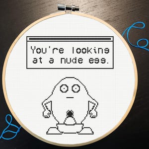 Può includere: Un telaio da ricamo a punto croce con un design umoristico. Il testo recita: "You're looking at a nude egg." Sotto il testo c'è un personaggio a forma di uovo dei cartoni animati. Il ricamo è in filo nero su tessuto bianco.