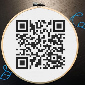 May include: A cross-stitch QR code in a wooden embroidery hoop. The QR code is black and white, stitched on white fabric. Blue thread is visible near the hoop.