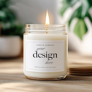 May include: A lit candle in a clear glass jar with a white label that reads "your design here." The candle sits on a wooden surface with a stack of wooden coasters nearby. Green plants are in the background.