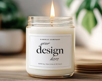 May include: A lit candle in a clear glass jar with a white label that reads "your design here." The candle sits on a wooden surface with a stack of wooden coasters nearby. Green plants are in the background.