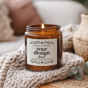 May include: A lit amber glass candle jar with a white label that reads "your design here" in a decorative font. The candle sits on a beige knitted blanket. The background includes a woven basket and decorative plants.