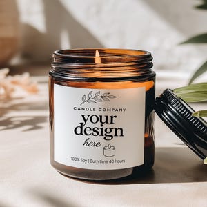 May include: A lit soy candle in a brown glass jar with a black lid. The label reads "Candle Company" and "your design here". The candle is on a light-colored surface with shadows and greenery.