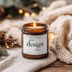 May include: A lit candle in a brown glass jar with a white label that reads "your design here." The candle is set on a cream-colored knitted blanket. The background includes pine needles, pine cones, and soft, out-of-focus lights.