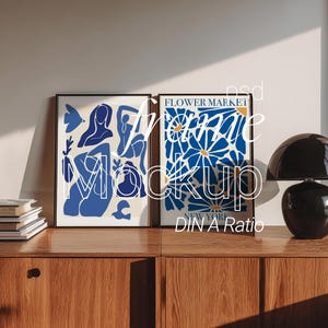Puede incluir: Dos láminas enmarcadas con marcos negros. Una presenta figuras abstractas azules sobre un fondo crema. La otra muestra un diseño floral azul con el texto "FLOWER MARKET NEW YORK". Una lámpara negra está sobre un mueble de madera.